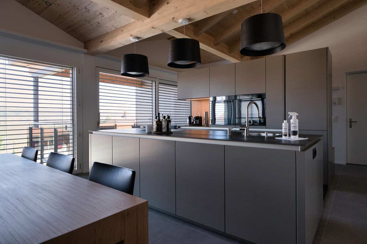 Offene Küche mit grauen, grifflosen Fronten, Kochinsel und schwarzen Designleuchten unter der Holzbalkendecke