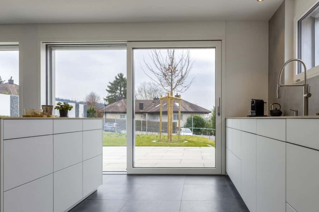 Helle weiße Küche mit großen Fenstern und Blick auf Garten und Terrasse.