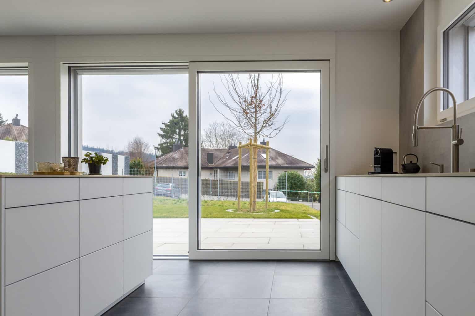 Helle weiße Küche mit großen Fenstern und Blick auf Garten und Terrasse.