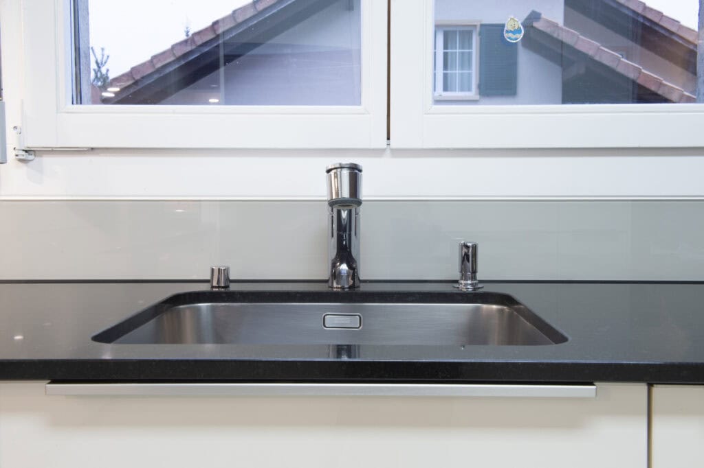 Moderne Küchenspüle mit schwarzem Granit-Design, Edelstahlarmatur und Fenster im Hintergrund.